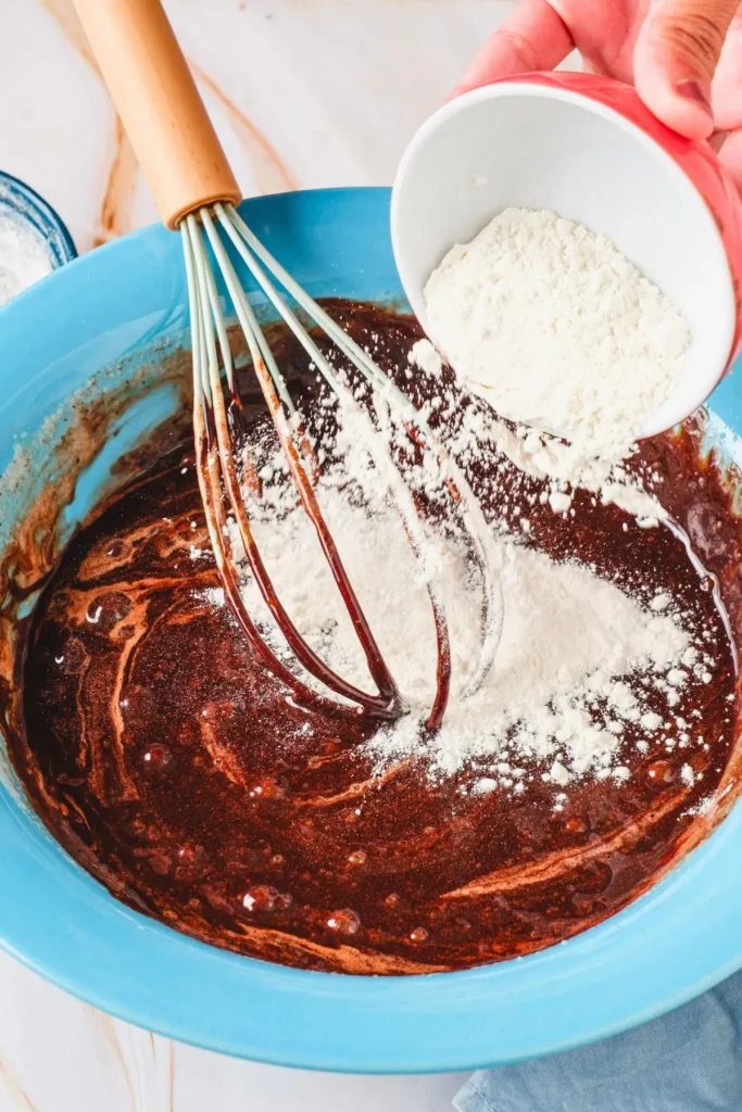 Hand mixing flour into chocolate batter with a whisk in a blue bowl, with a red bowl of flour nearby.