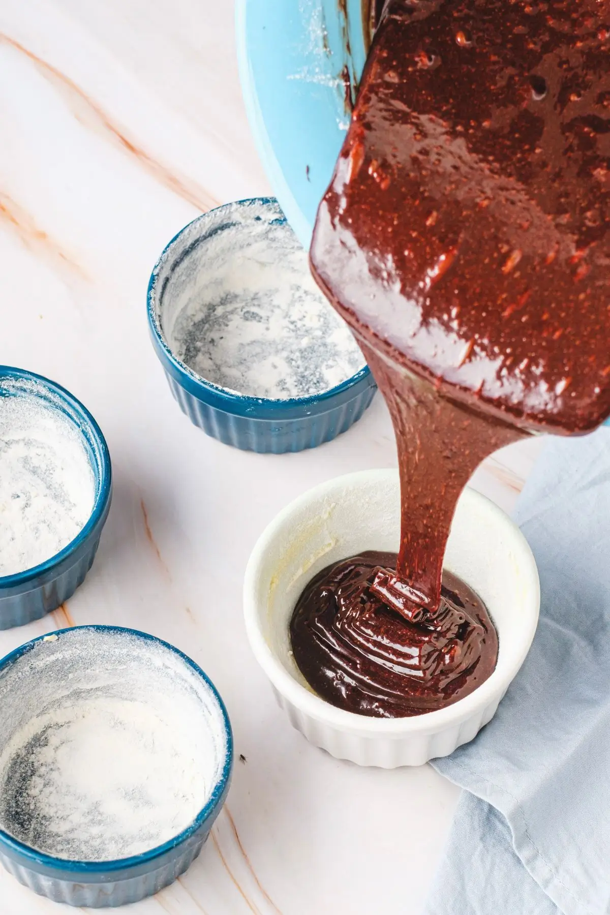 Molten Lava Chocolate Cake Chocolate batter being poured into a white ramekin, surrounded by floured blue ramekins on a marble surface.