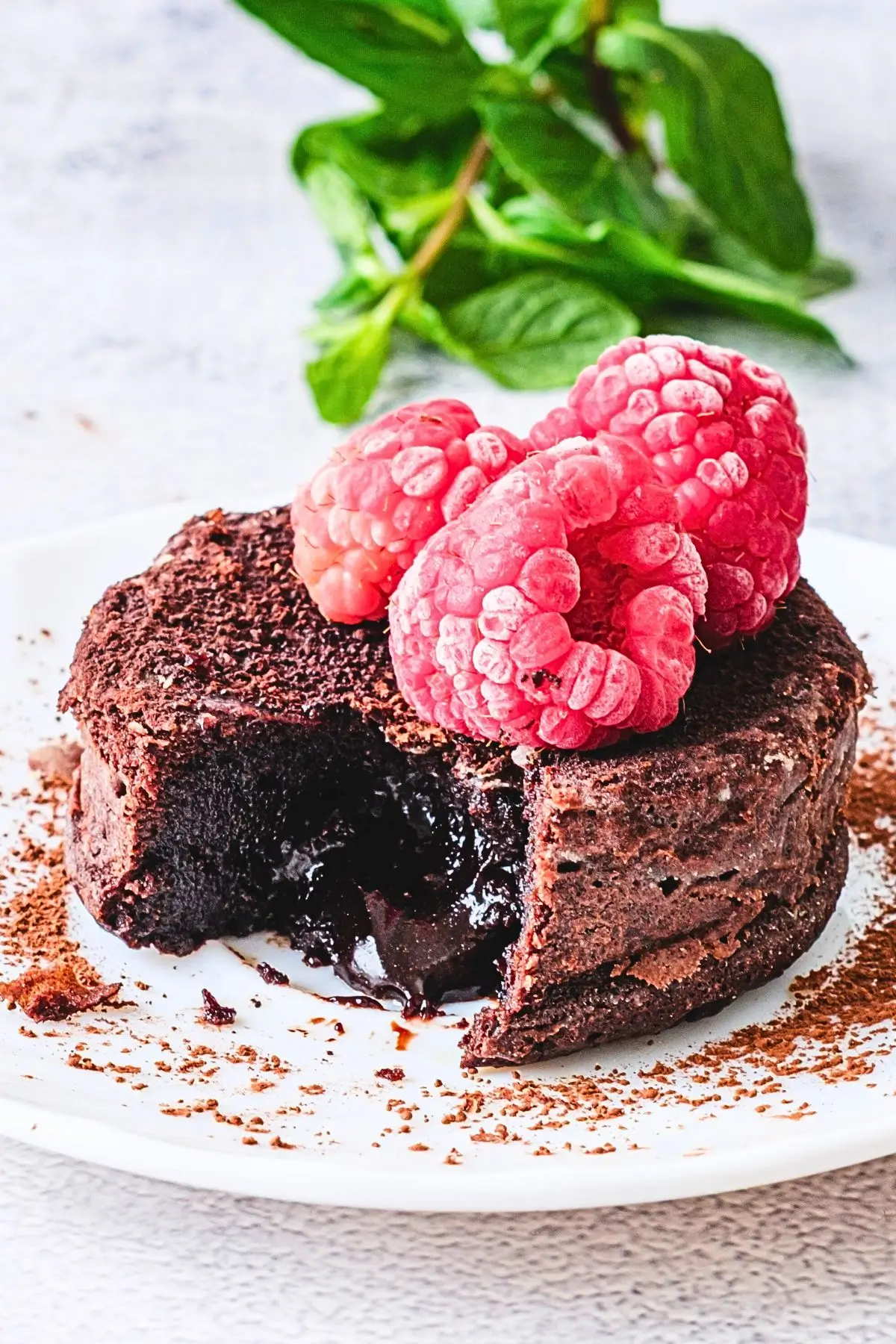 Molten Lava Chocolate Cake Molten lava chocolate cake on a white plate, cut open with flowing chocolate, topped with three raspberries and mint leaves, garnished with cocoa powder.