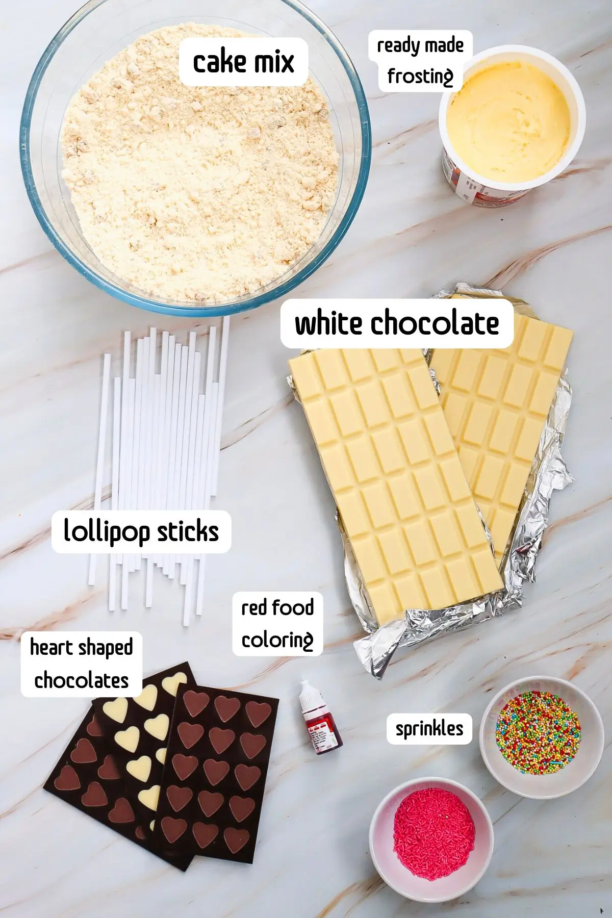 Flat lay of cake pop ingredients: cake mix, frosting, white chocolate, sticks, red food coloring, heart-shaped chocolates.