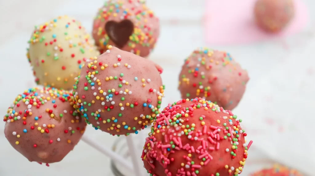 Colorful sprinkle-covered cake pops on sticks with pink, red, and white icing, fun party dessert