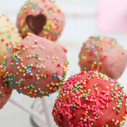 Colorful sprinkle-covered cake pops on sticks with pink, red, and white icing, fun party dessert