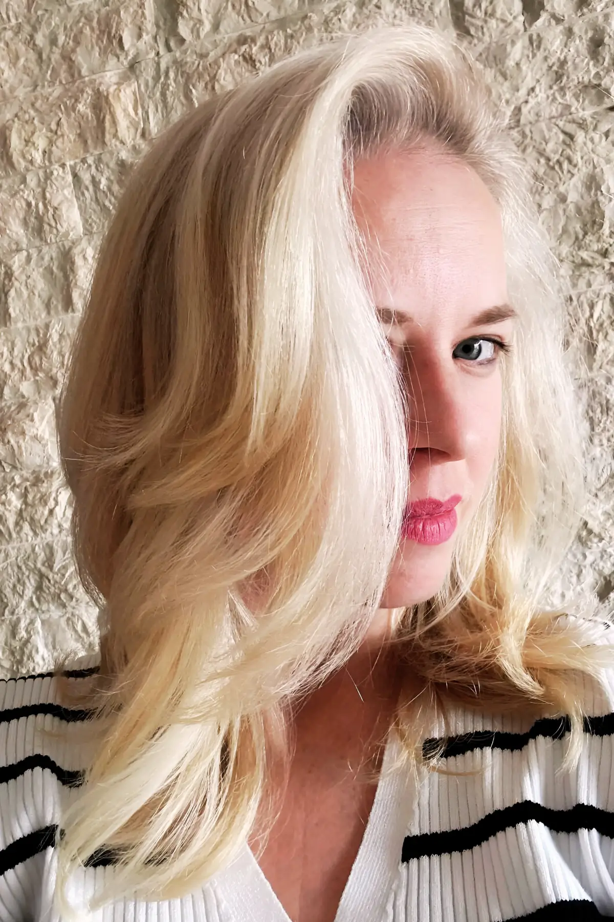 Close-up of blonde woman with layered wavy hair and pink lipstick against stone wall
