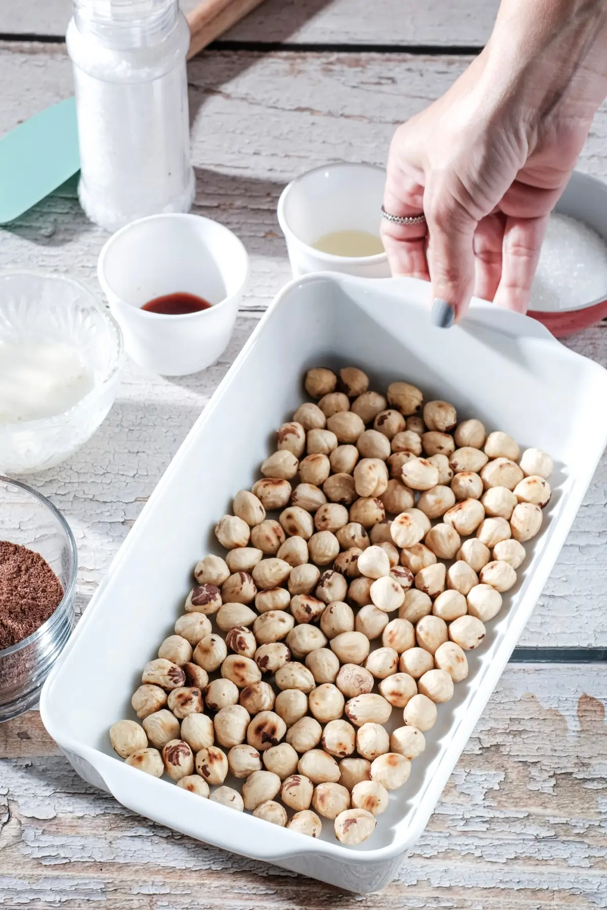 Hand holding a baking dish of raw hazelnuts with bowls of ingredients on rustic table