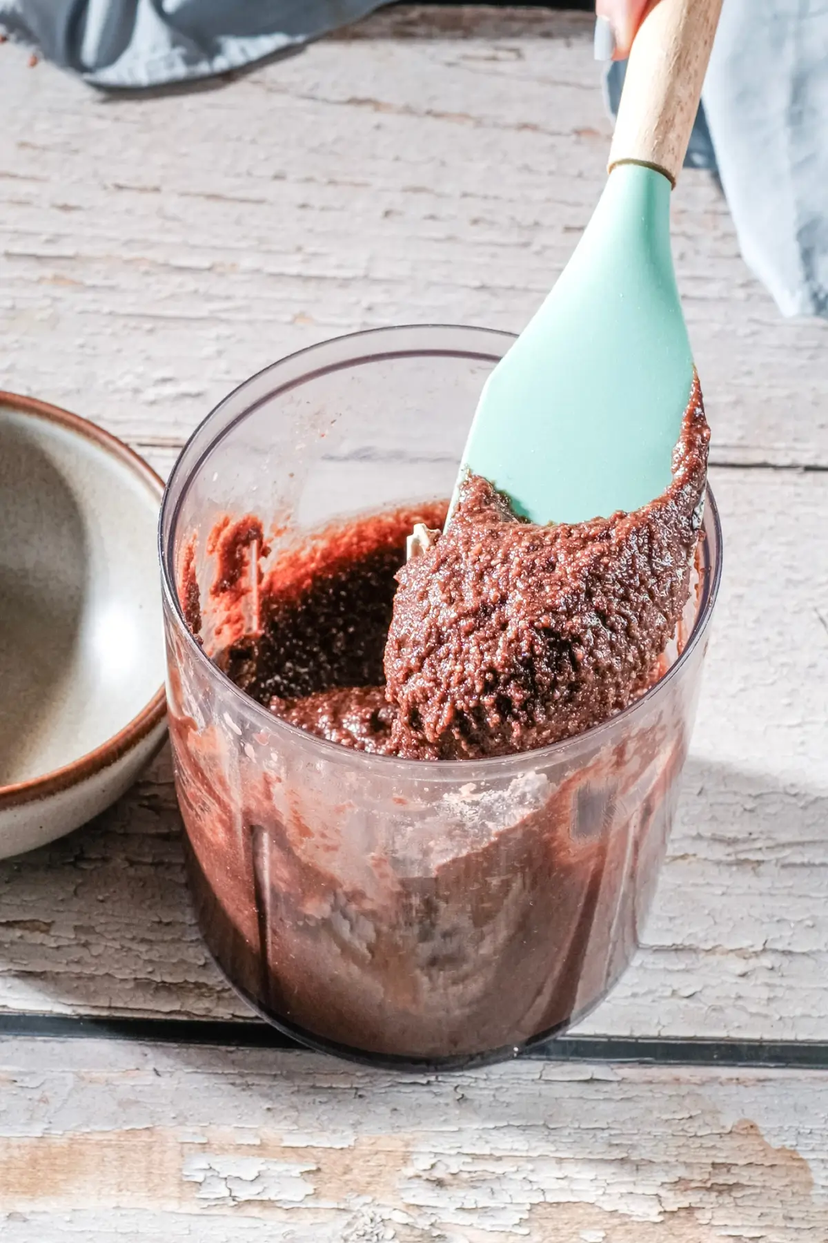 Thick chocolate batter in a food processor cup, scooped with a mint spatula on a rustic table