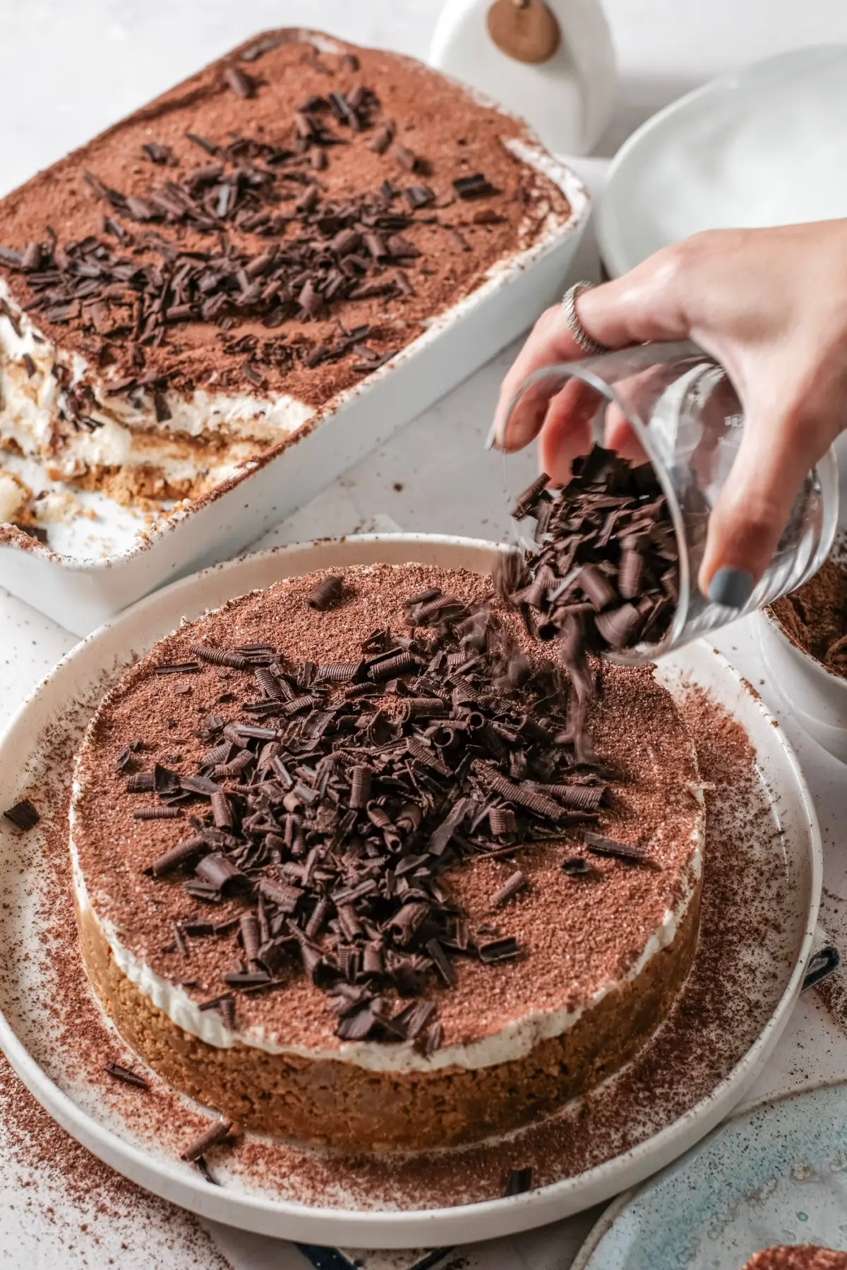 Hand sprinkling chocolate shavings over tiramisu chceesecake cake with cocoa powder topping