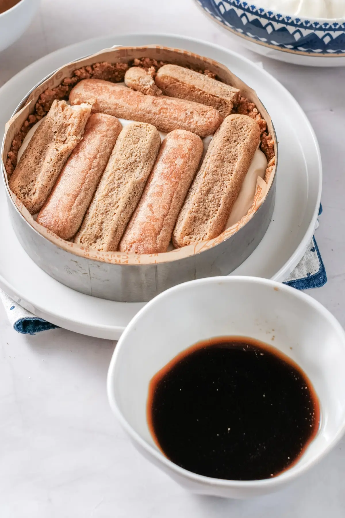 Tiramisu assembly with soaked ladyfingers over mascarpone in a ring mold, beside a bowl of espresso