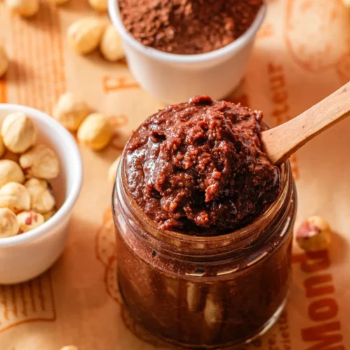 Jar of homemade chocolate hazelnut spread with wooden spoon, cocoa powder and raw hazelnuts.