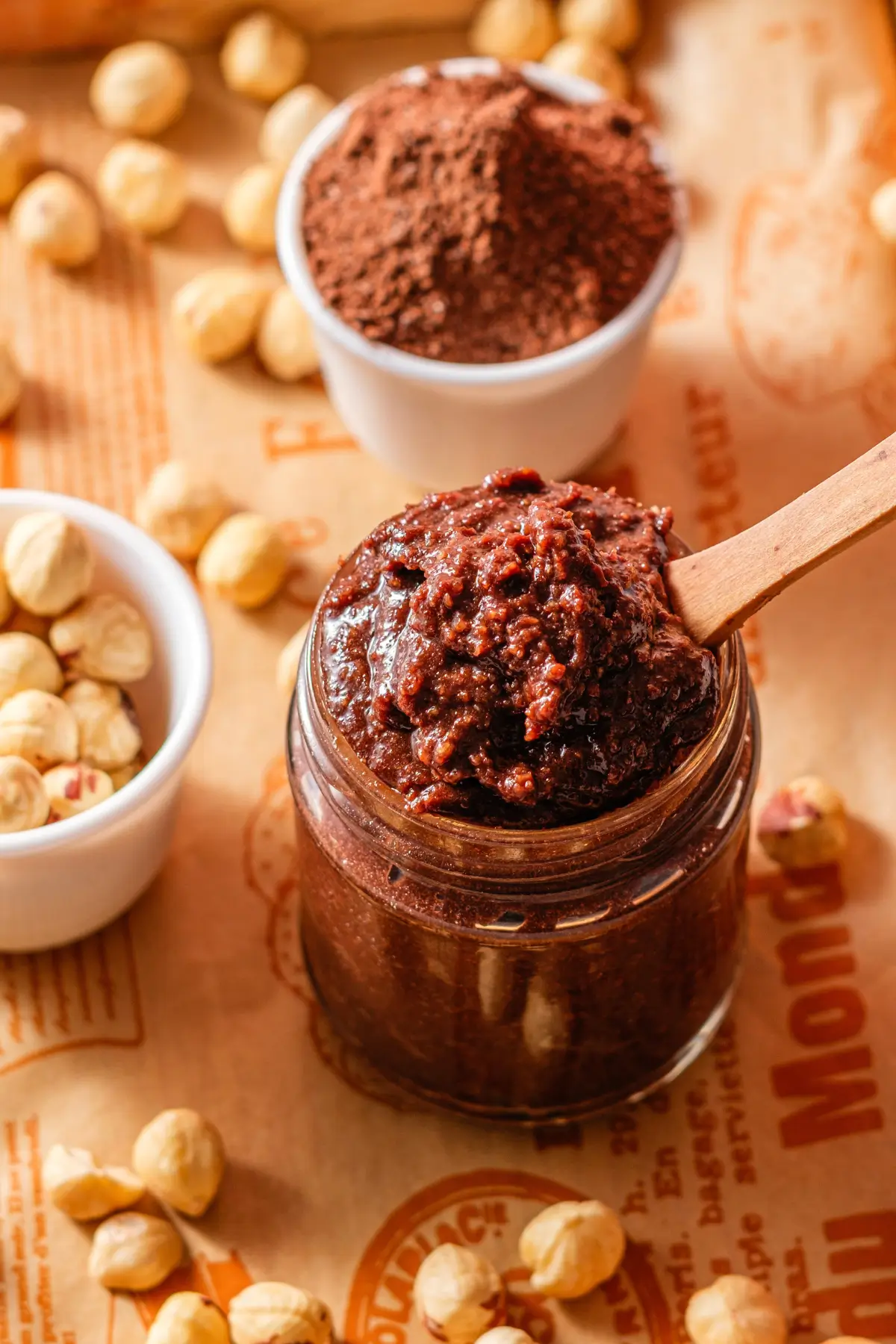 Jar of homemade chocolate hazelnut spread with wooden spoon, cocoa powder and raw hazelnuts.