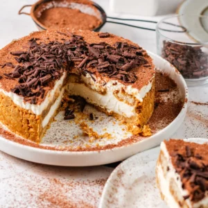 Creamy tiramisu cheesecake topped with chocolate shavings and cocoa, with a slice cut on a white plate