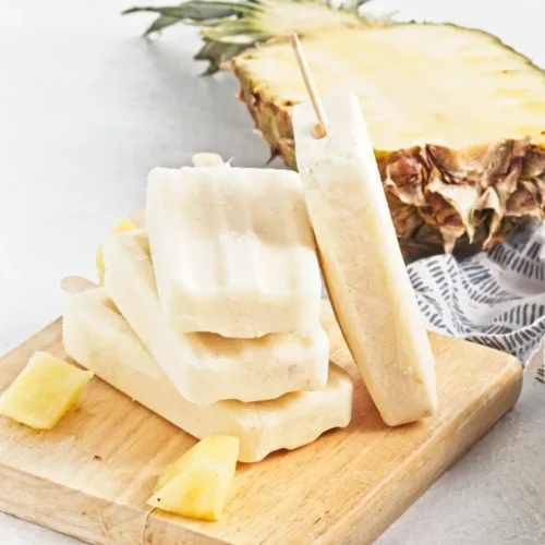 Creamy pina colada popsicles stacked on a wooden board with pineapple