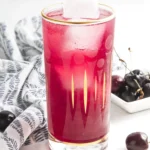 Tall glass of homemade cherry vanilla soda with ice and fresh cherries nearby.