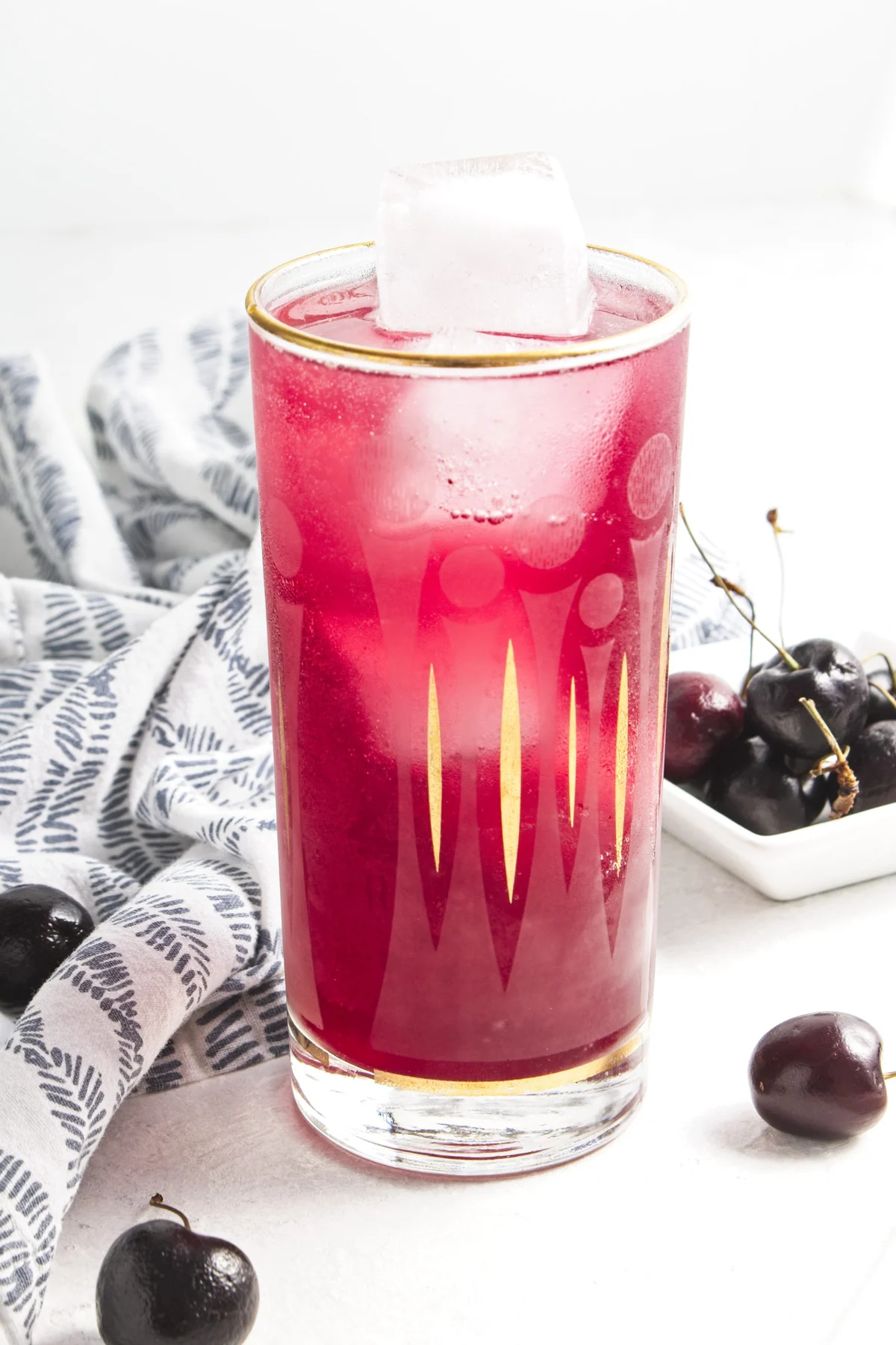 Tall glass of homemade cherry vanilla soda with ice and fresh cherries nearby.