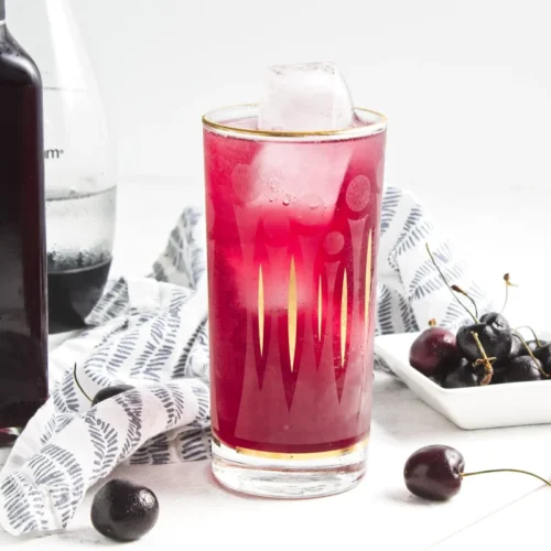 A glass of bright pink cherry vanilla soda with ice, a bottle of cherry syrup, and fresh cherries.
