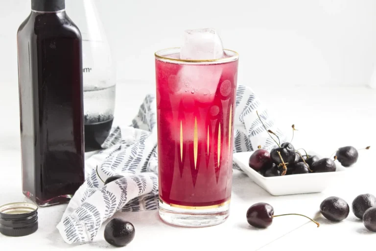 A glass of bright pink cherry vanilla soda with ice, a bottle of cherry syrup, and fresh cherries.