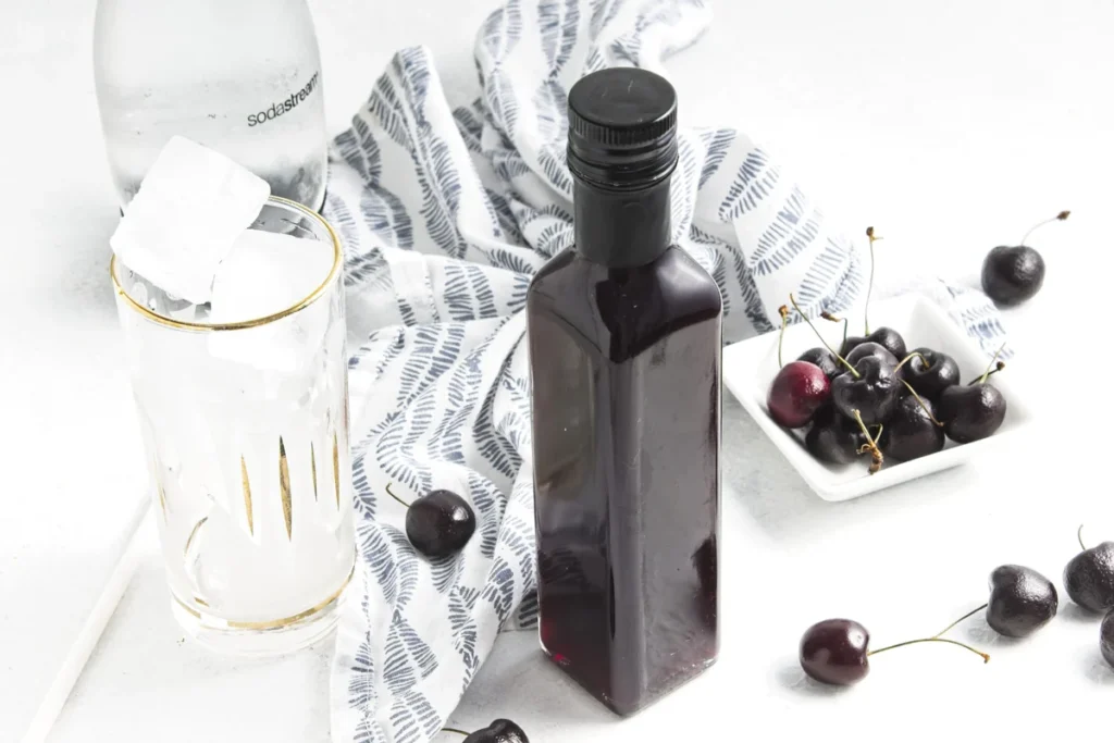 Bottle of cherry vanilla syrup with a glass, soda water, and fresh cherries.