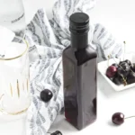 Bottle of cherry vanilla syrup with a glass, soda water, and fresh cherries.