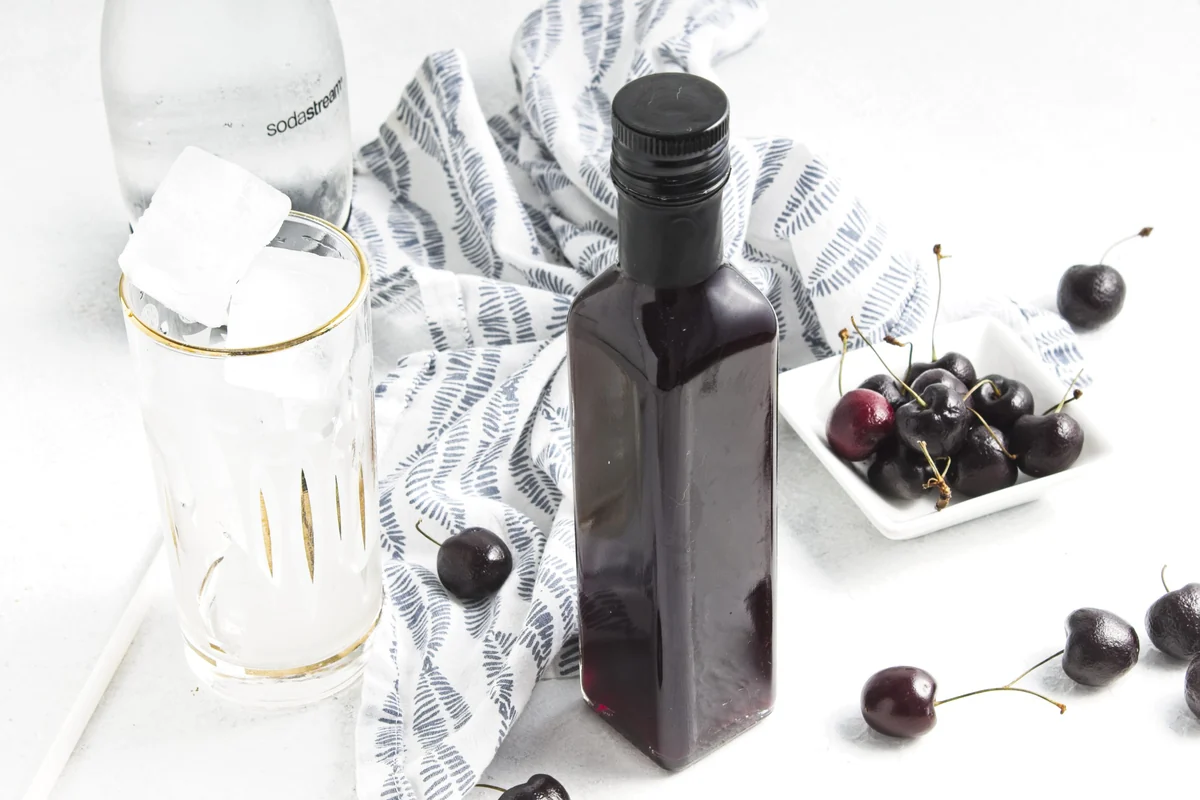 Bottle of cherry vanilla syrup with a glass, soda water, and fresh cherries.