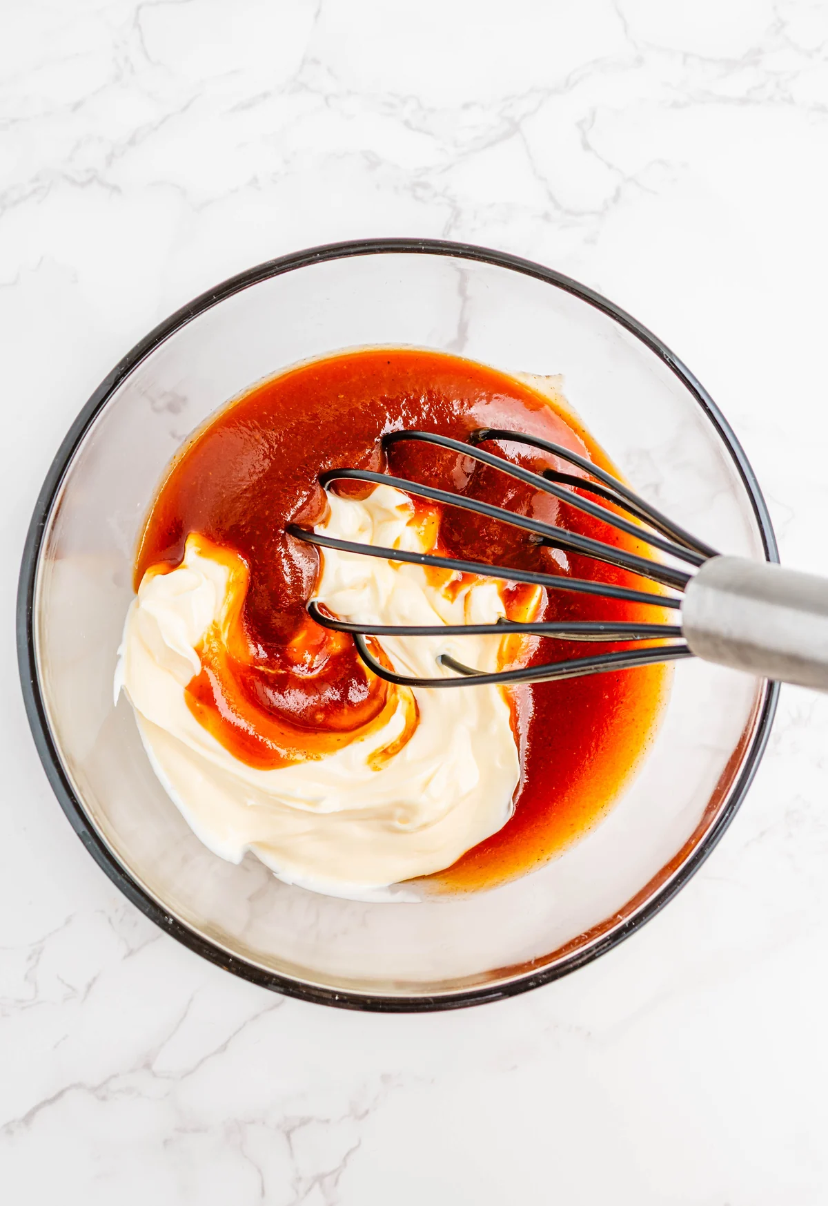 A whisk beginning to mix mayonnaise and BBQ sauce in a glass bowl.