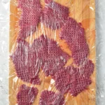 Pounded venison pieces on a cutting board under plastic wrap