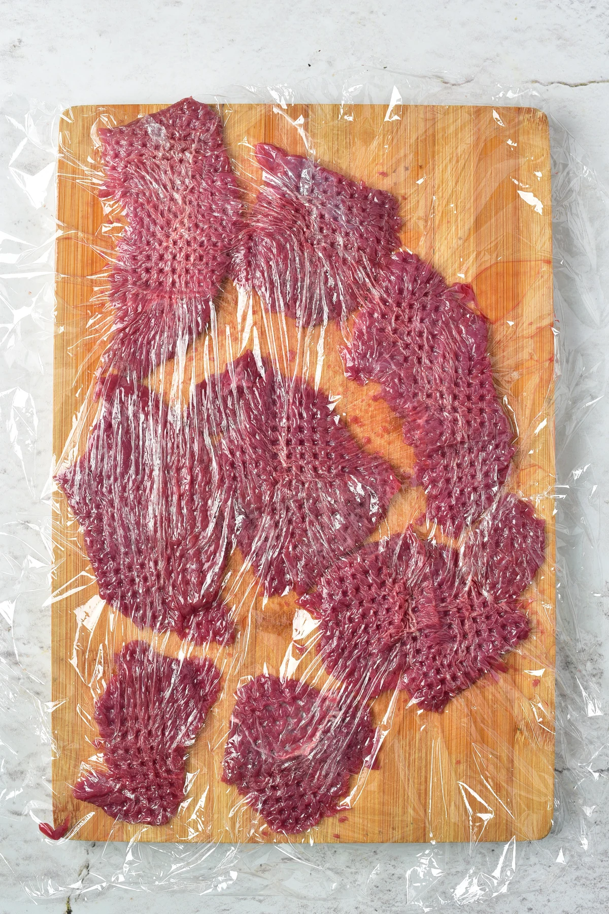 Pounded venison pieces on a cutting board under plastic wrap