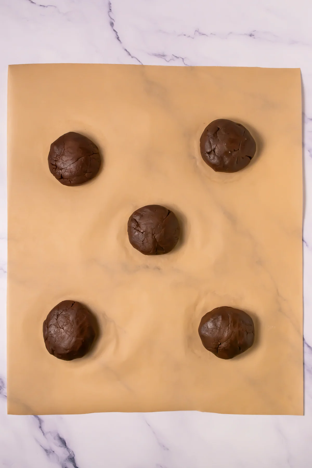 Chocolate cookie dough balls sealed around marshmallow filling on parchment paper.