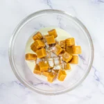 Crumbl Copycat Twix Cookies With Caramel and Toffee Caramel candies and heavy cream ready to melt in a glass bowl.