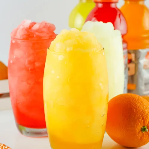 Trio of colorful slushie drinks in tall glasses with crushed ice, orange fruit and bottles in background