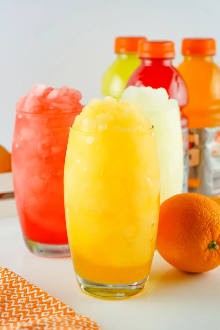 Trio of colorful slushie drinks in tall glasses with crushed ice, orange fruit and bottles in background