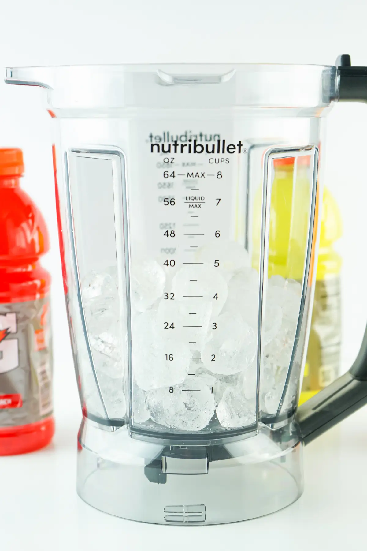 Nutribullet blender jar filled with ice, clear measurement markings, red and yellow sports drink bottles behind.