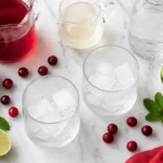 Glasses filled with ice beside cranberry juice, lime, mint, and sparkling water.