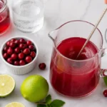 Cranberry juice, lime juice, and simple syrup being stirred together in a glass pitcher.