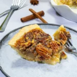 Slice of bourbon pecan pie served on a plate with cinnamon and bourbon in the background.