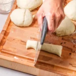 Pretzel dough rope being cut into bite-sized pieces on a wooden board.