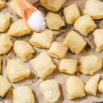 Coarse salt being sprinkled over pretzel bites on a parchment-lined baking sheet.