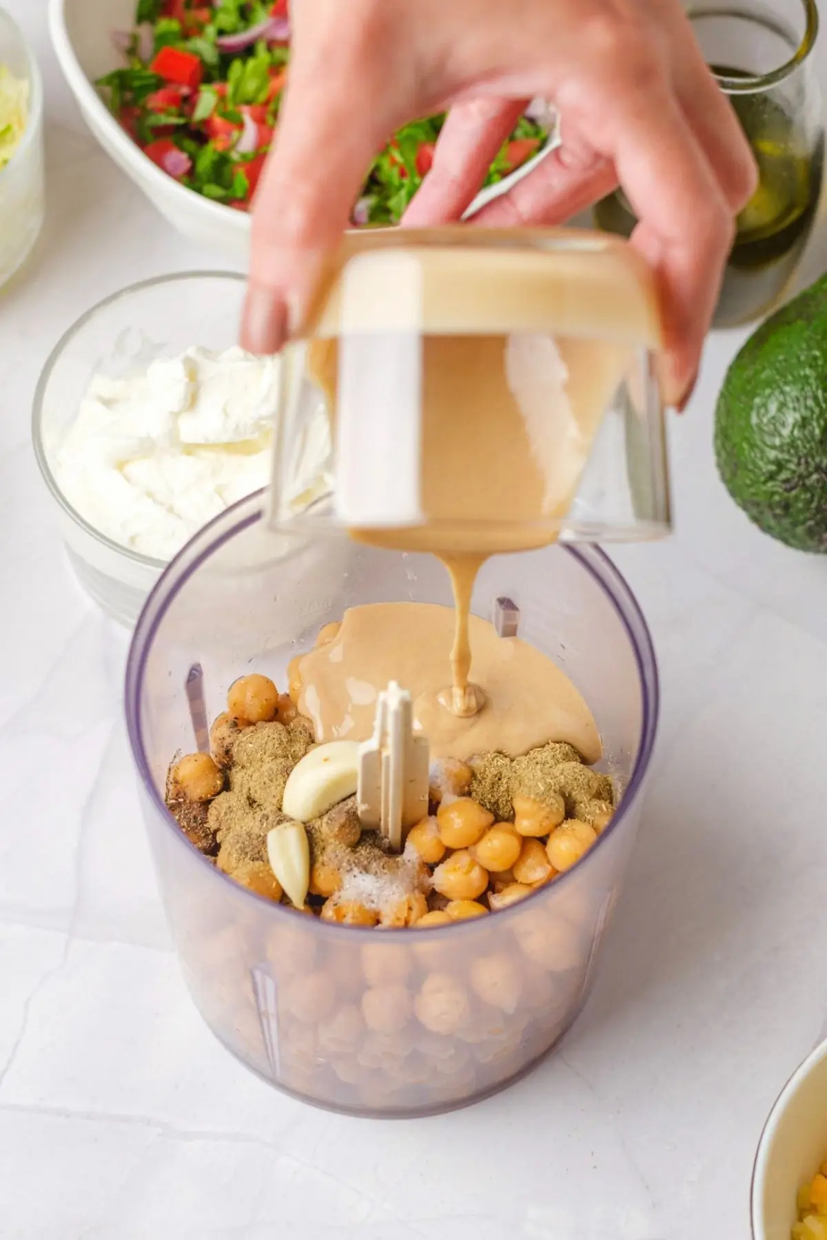 Pouring drained chickpeas into a food processor, then adding tahini over the chickpeas and garlic