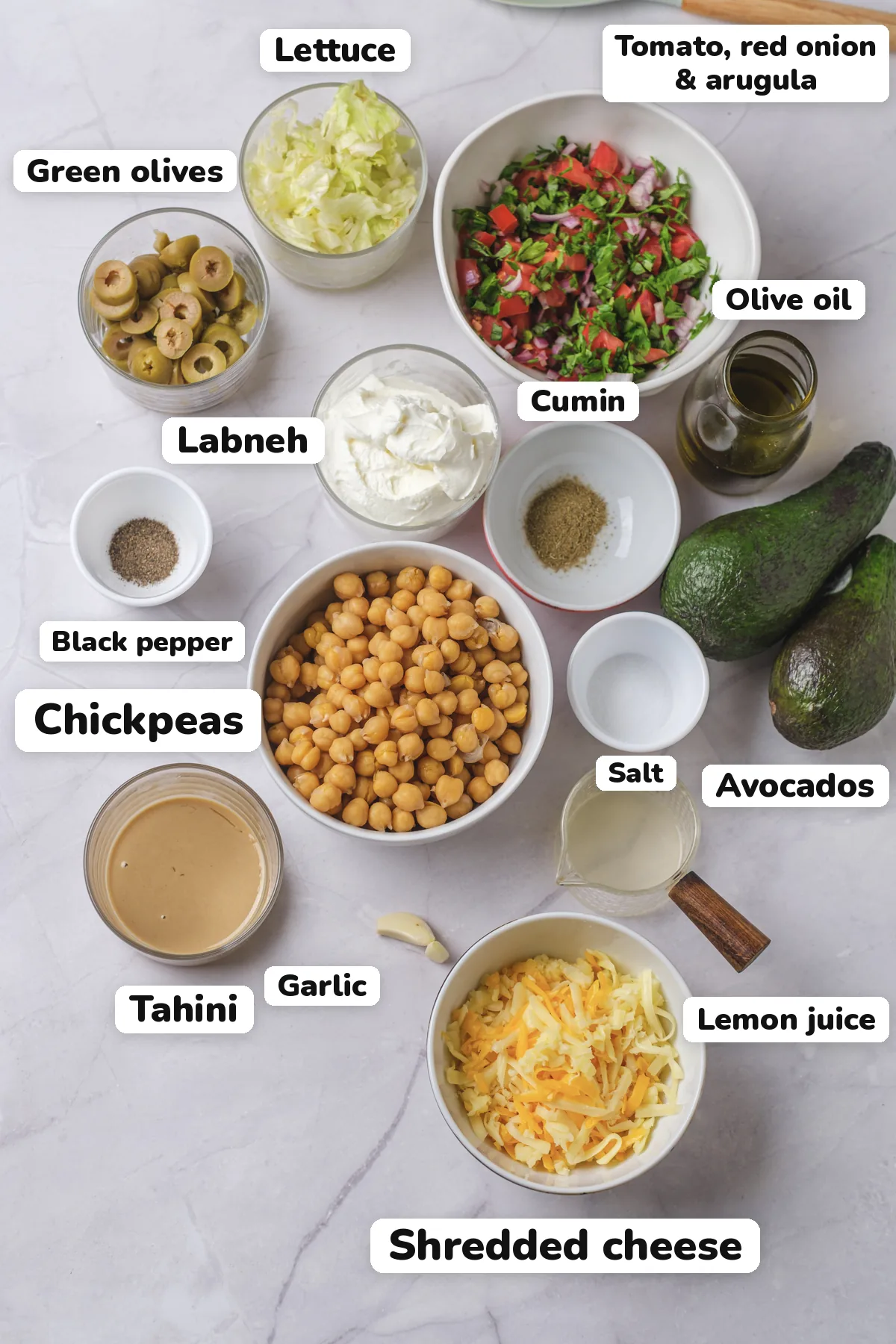 Labeled ingredients for 7 layer dip: chickpeas, tahini, garlic, cumin, lemon juice, olive oil, salt, black pepper, avocados, labneh, tomato, red onion and chopped arugula, shredded cheddar and Monterey Jack, lettuce and green olives