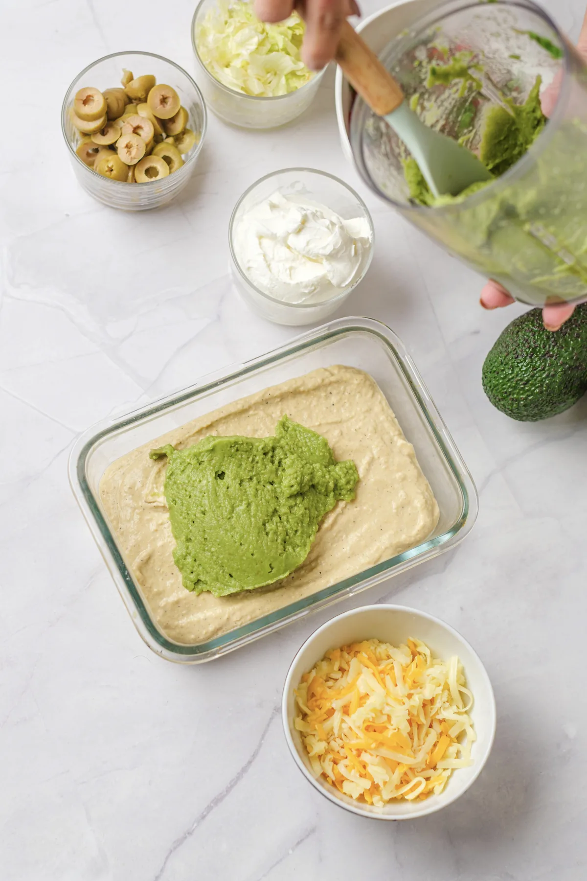 Spooning blended avocado over the hummus layer in a glass dish