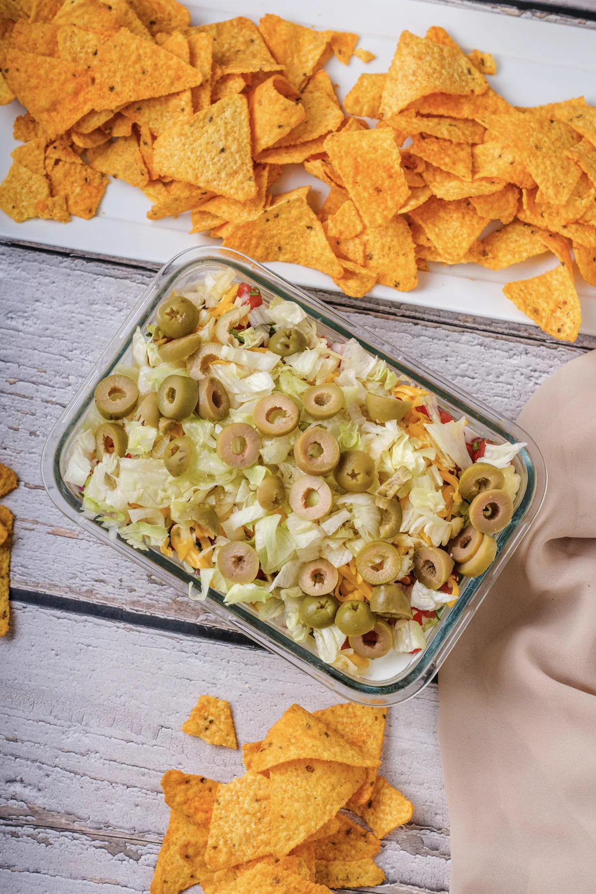 Overhead view of 7 layer dip topped with lettuce and sliced green olives in a glass dish, with tortilla chips and a beige napkin on a white wood table
