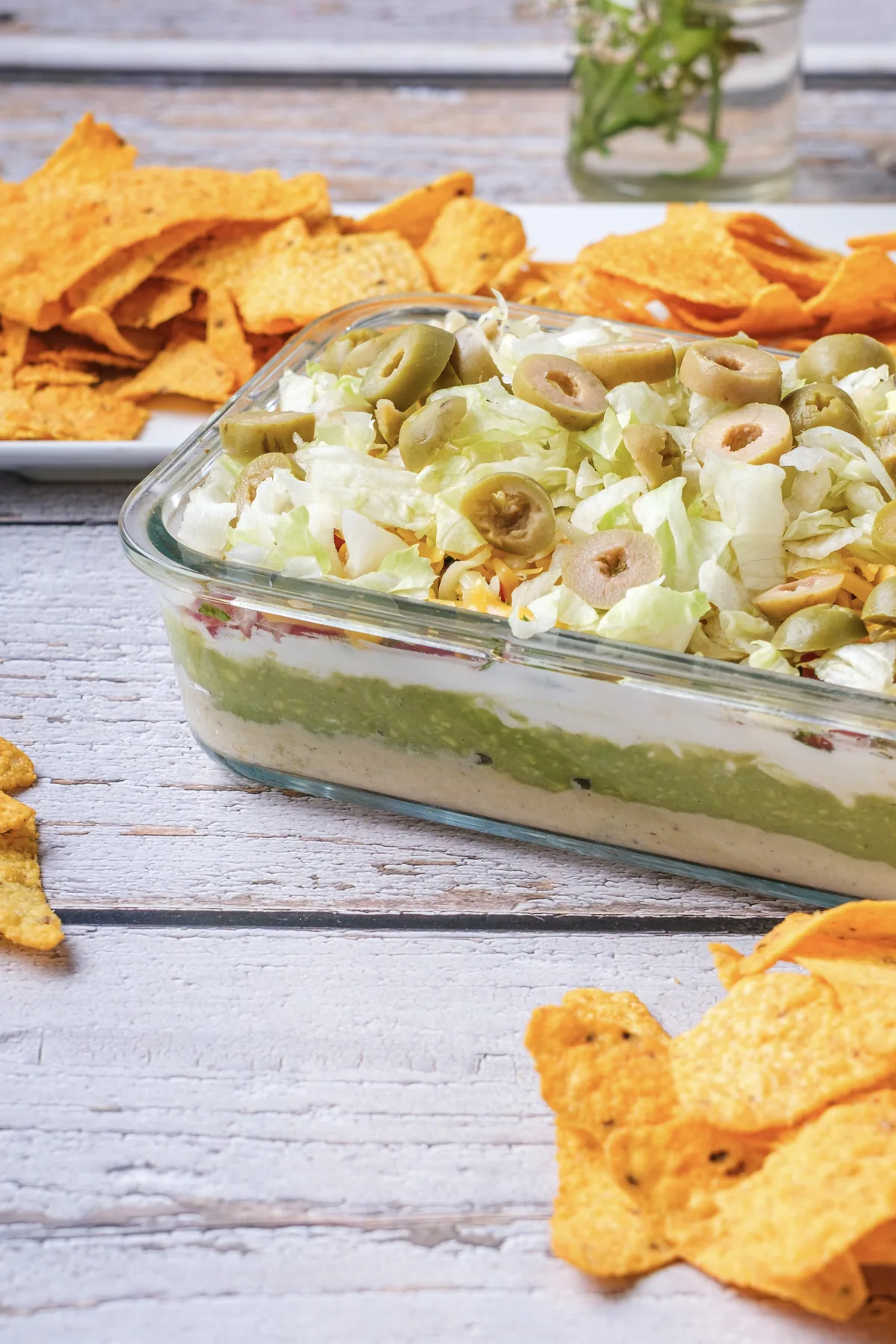 7 layer dip in a glass dish seen from the side, with hummus, avocado and labneh layers under tomato, cheese, lettuce and green olives, next to tortilla chips