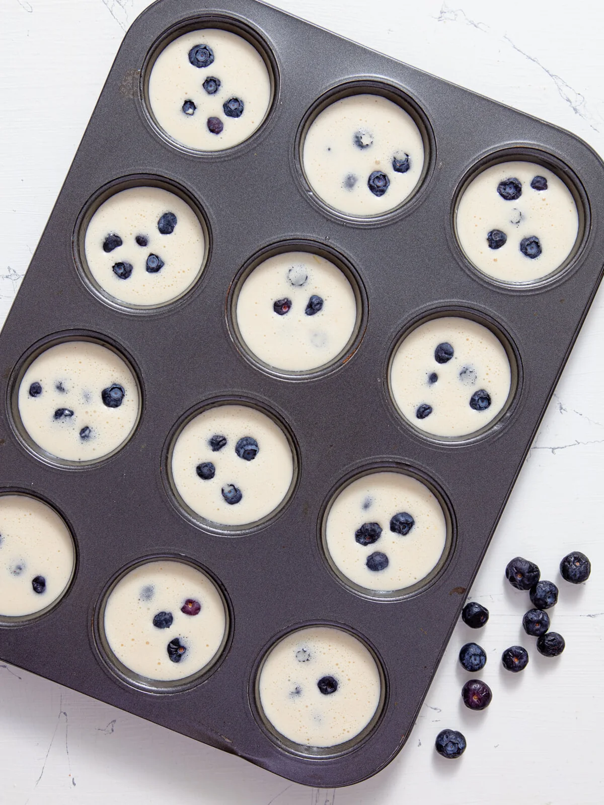 Muffin cups filled with thin popover batter and fresh blueberries before baking.