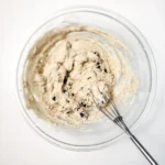 Thick blueberry muffin batter in a mixing bowl with a whisk.