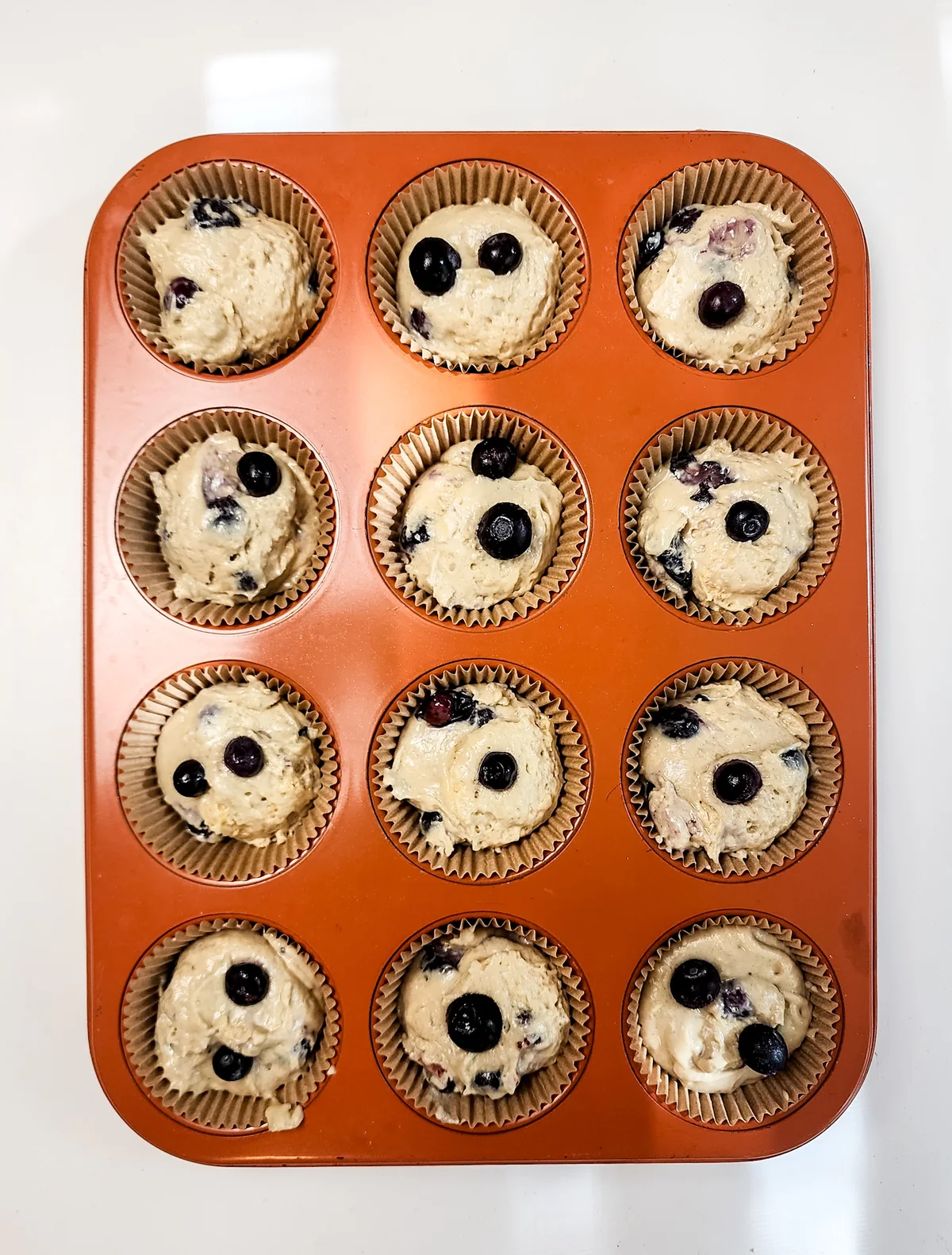 Twelve paper-lined muffin cups filled with blueberry muffin batter before baking.