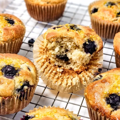 Blueberry muffins with golden tops on a cooling rack, including one split open to show the crumb.