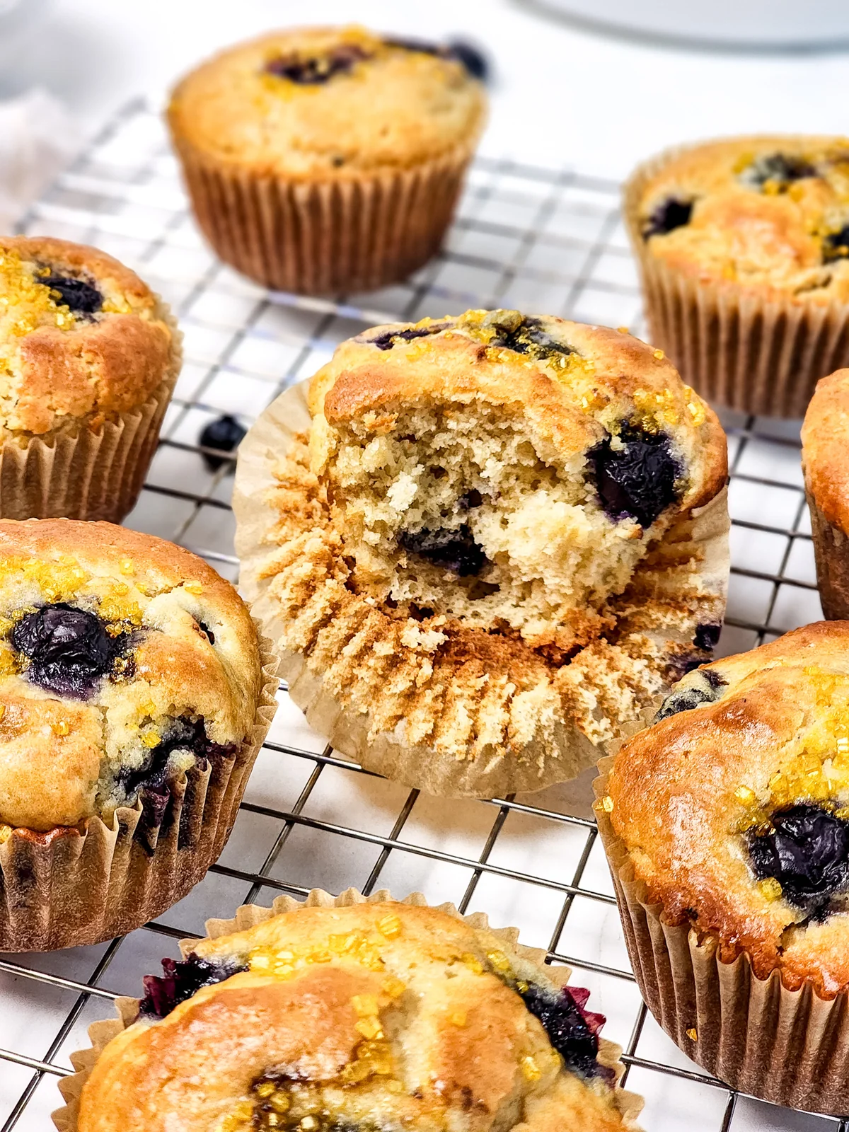 Blueberry muffins with golden tops on a cooling rack, including one split open to show the crumb.