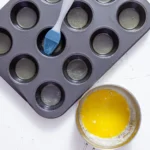 A standard muffin tin being coated with melted butter using a pastry brush.