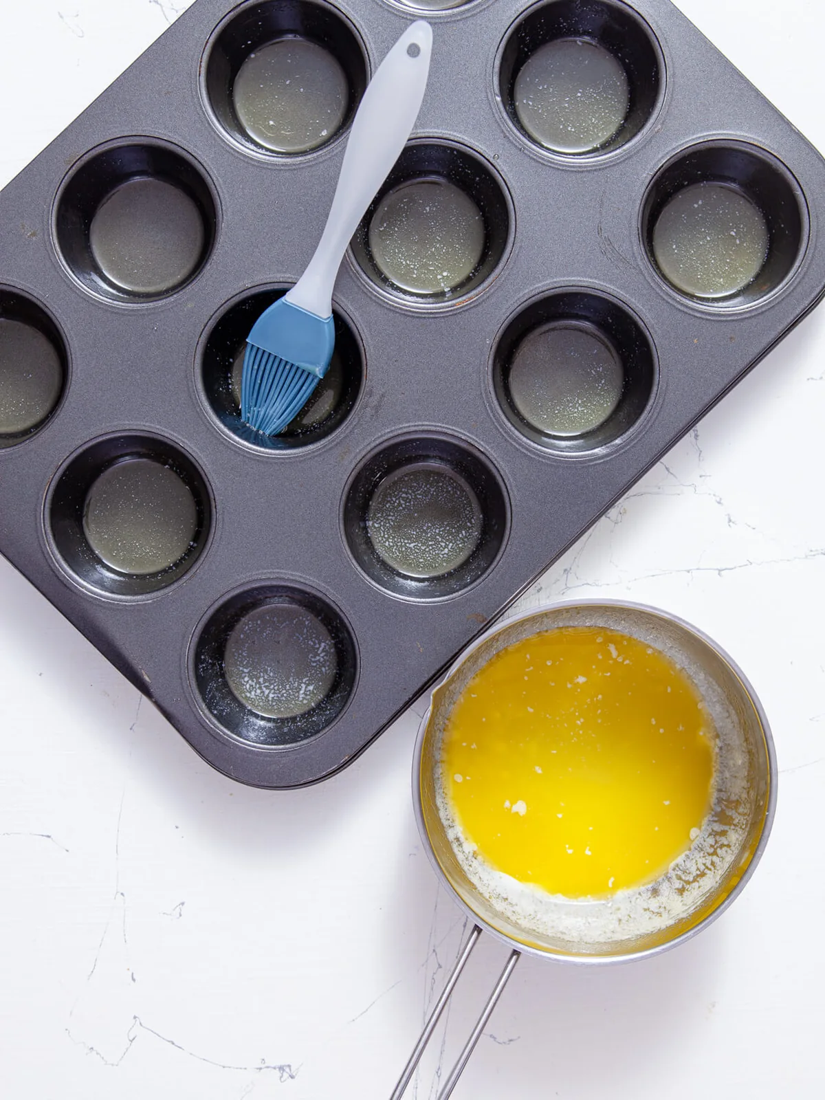 A standard muffin tin being coated with melted butter using a pastry brush.