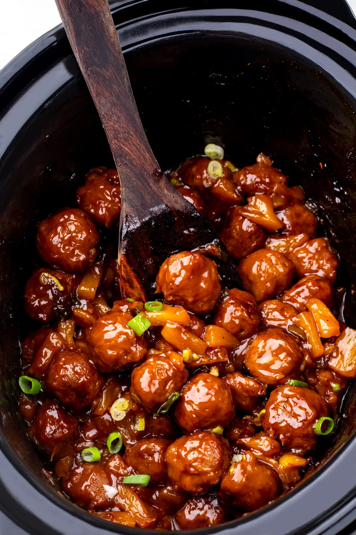 A wooden spoon lifting cooked Hawaiian meatballs in thick BBQ pineapple sauce from a slow cooker