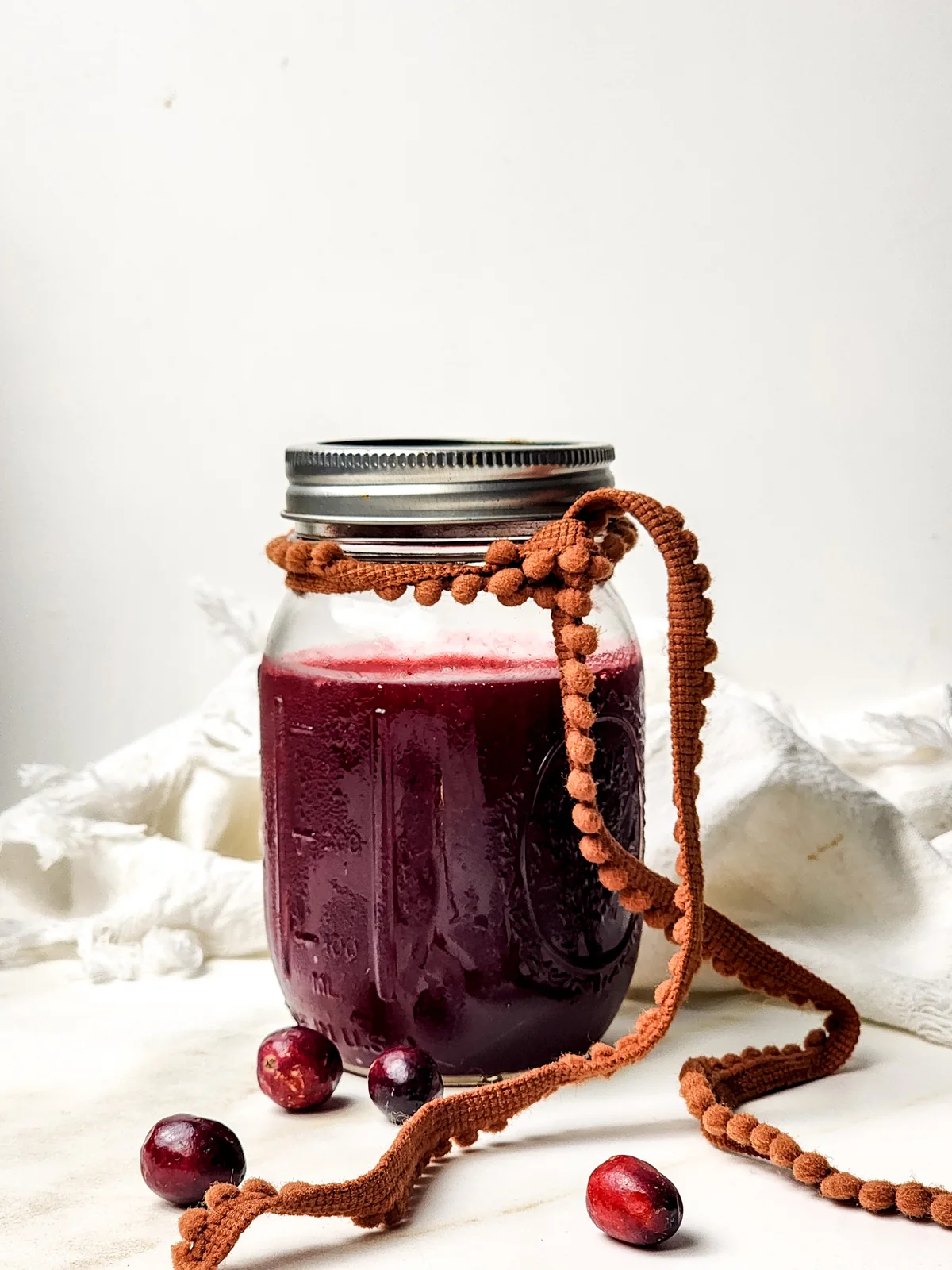 Glass jar of cranberry sauce with a silver lid and orange ribbon, surrounded by fresh cranberries.