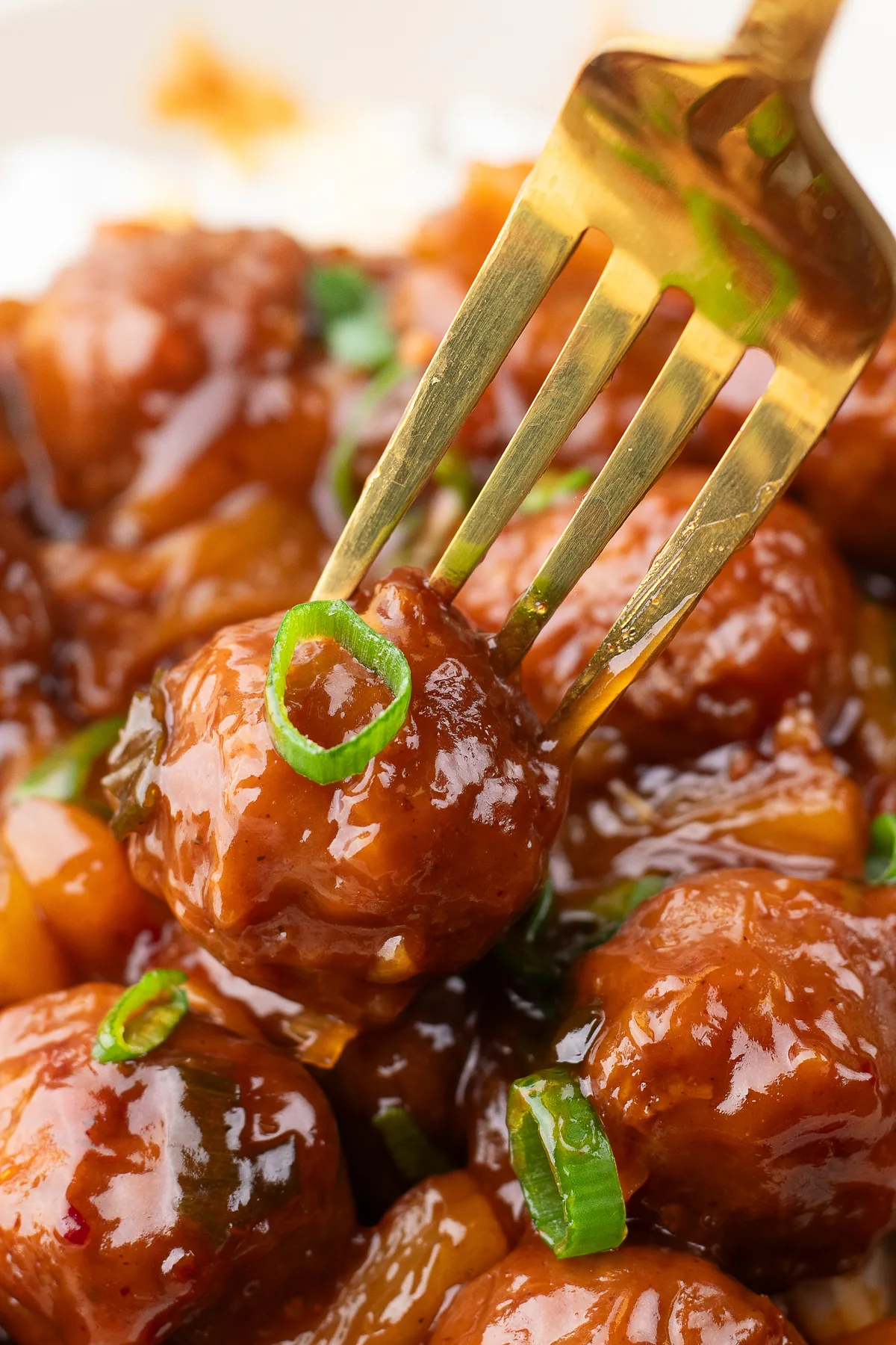 A gold fork lifting a spicy Hawaiian meatball with BBQ sauce, pineapple and a green onion slice